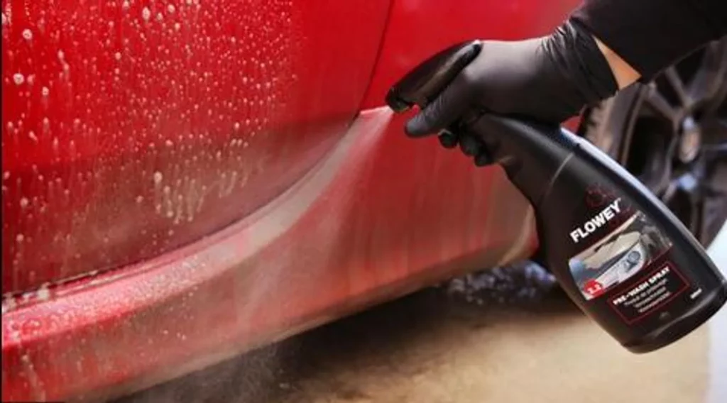 Ein glänzendes Auto wird mit Flowey Pre-Wash Spray behandelt. Das Produkt wird auf die Oberfläche gespritzt. Es befinden sich Schaumreste auf der Karosserie. Das Auto kann somit zuhause gewaschen werden.