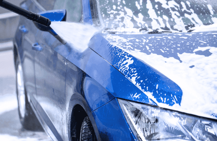 Ein glänzendes Auto wird mit einem Hochdruckreiniger gewaschen, während Wasserstrahlen über die Oberfläche spritzen. Es befinden sich schaumreste auf der Karosserie. Das Auto kann somit zuhause gewaschen werden.